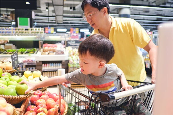 Asian toddler sitting in a grocery cart reaches out for an apple at the supermarket while his dad looks on.