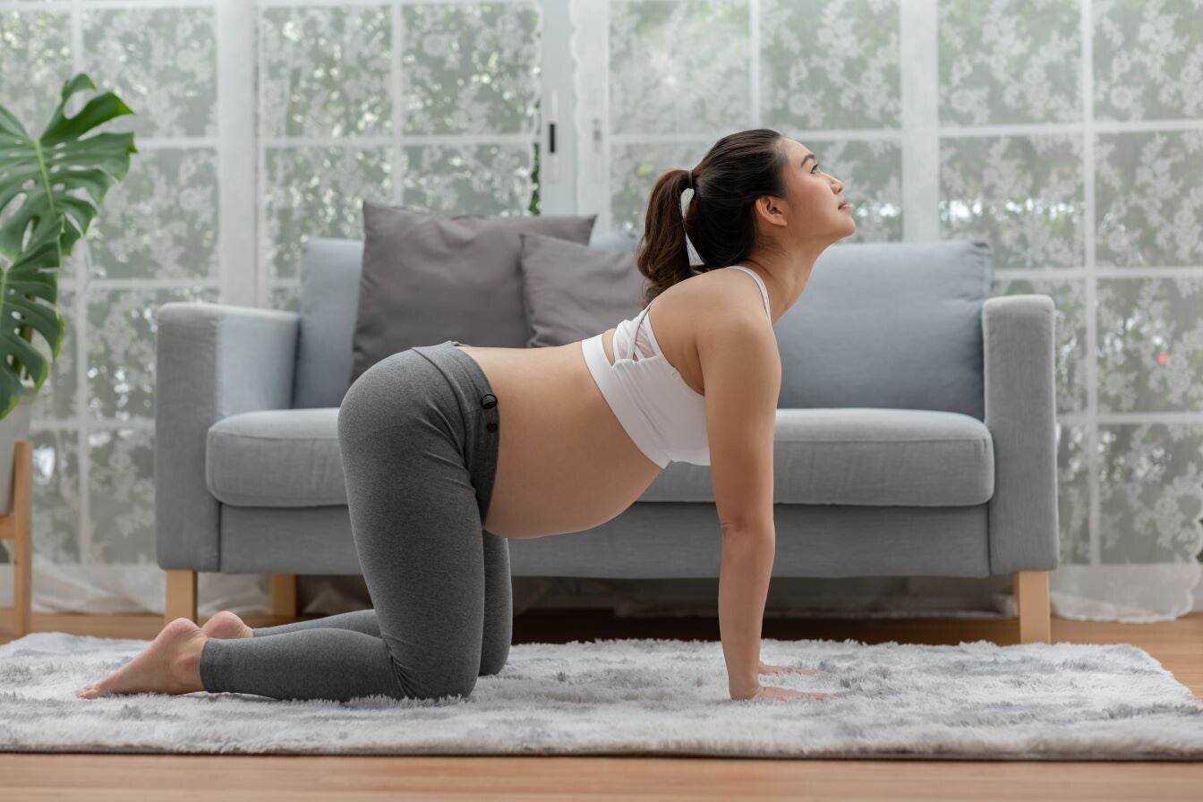 Pregnant Asian woman doing stretches on carpet in front of gray sofa.