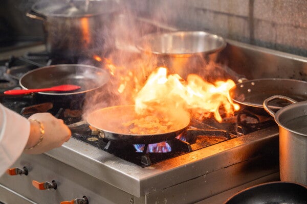Photo of a cropped hand holding a pan with meat on fire over a stove.
