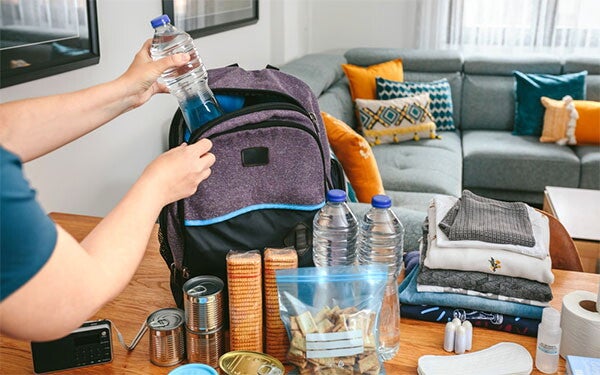 Close-up of a woman’s hand putting a water bottle in a backpack in the living room, with other emergency supplies on the table.