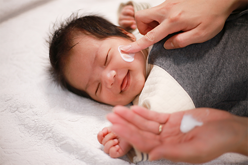 Close-up photo of a mother’s hands applying lotion to a smiling baby’s cheeks
