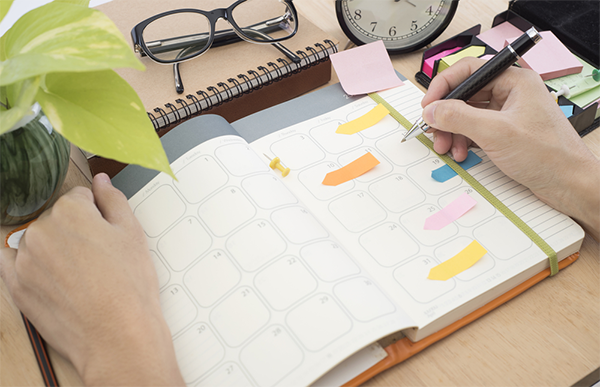 Close-up pic of a hand holding a pen to mark the 2026 Philippine Holidays on a planner beside a plant, eyeglasses, and clock