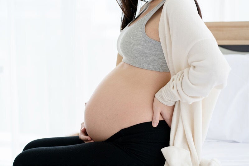 Close-up shot of a pregnant woman’s torso as she sits in bed and holds her belly with one hand, while the other supports her lower back.