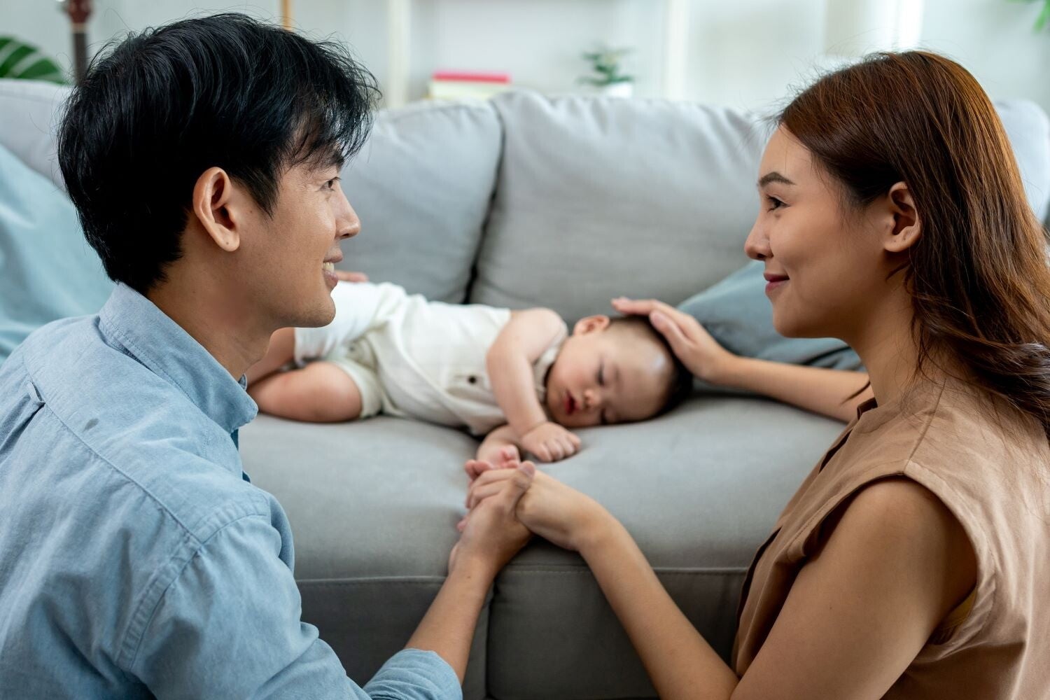 Young Asian couple holds each other hands in front of their sleeping baby on the sofa.