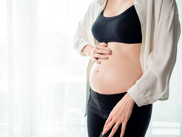 Cropped photo of a 14 weeks pregnant woman’s torso touching her belly against white curtains.