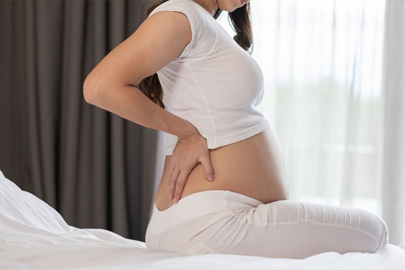 Cropped photo of a pregnant Asian woman’s torso seated on bed, with hands placed on lower back.