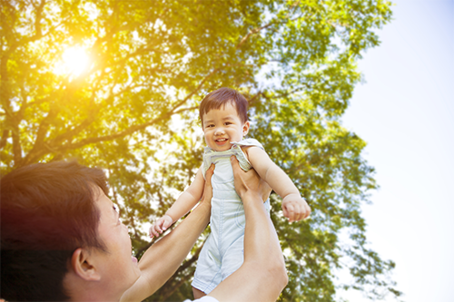 Cropped shot of an Asian father lifting his toddler boy outdoors with the sun and trees in the background