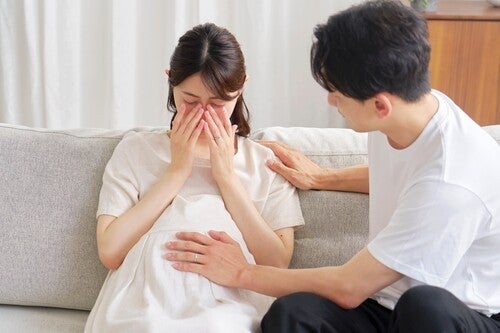 A pregnant Asian woman is crying on the sofa while her husband comforts her