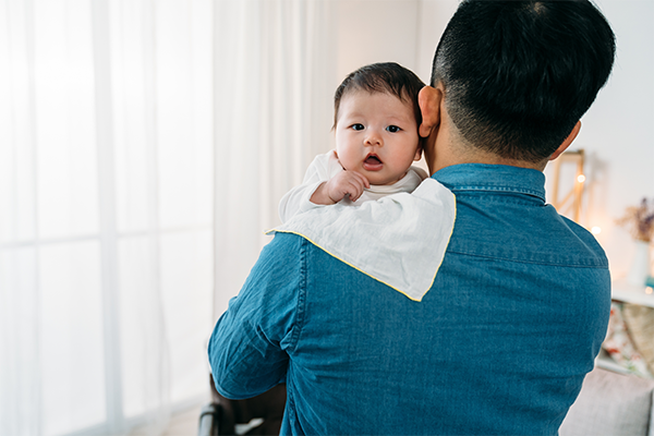 Photo of a black-haired father’s back carrying his infant to burp him.