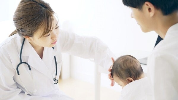 A female Asian doctor examining a baby’s head and hair while his dad holds him.