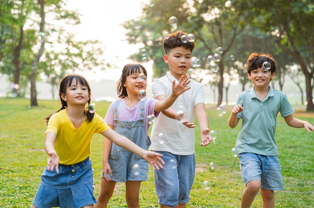 Four Asian children of different genders and heights run after bubbles at a park.