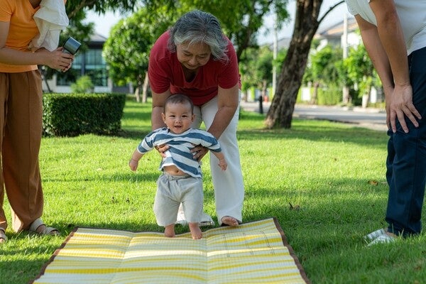 Grandmother helping her grandchild learn to walk in the park while parents watch on.