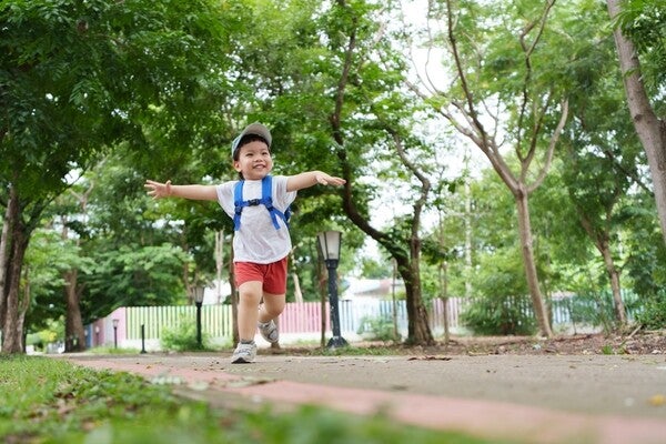 A happy Asian boy running outdoors with a cap and backpack.