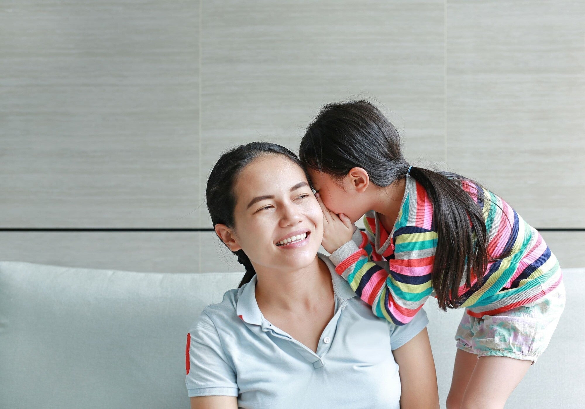 Asian girl stands up on the sofa to whisper in her mom's ear.  