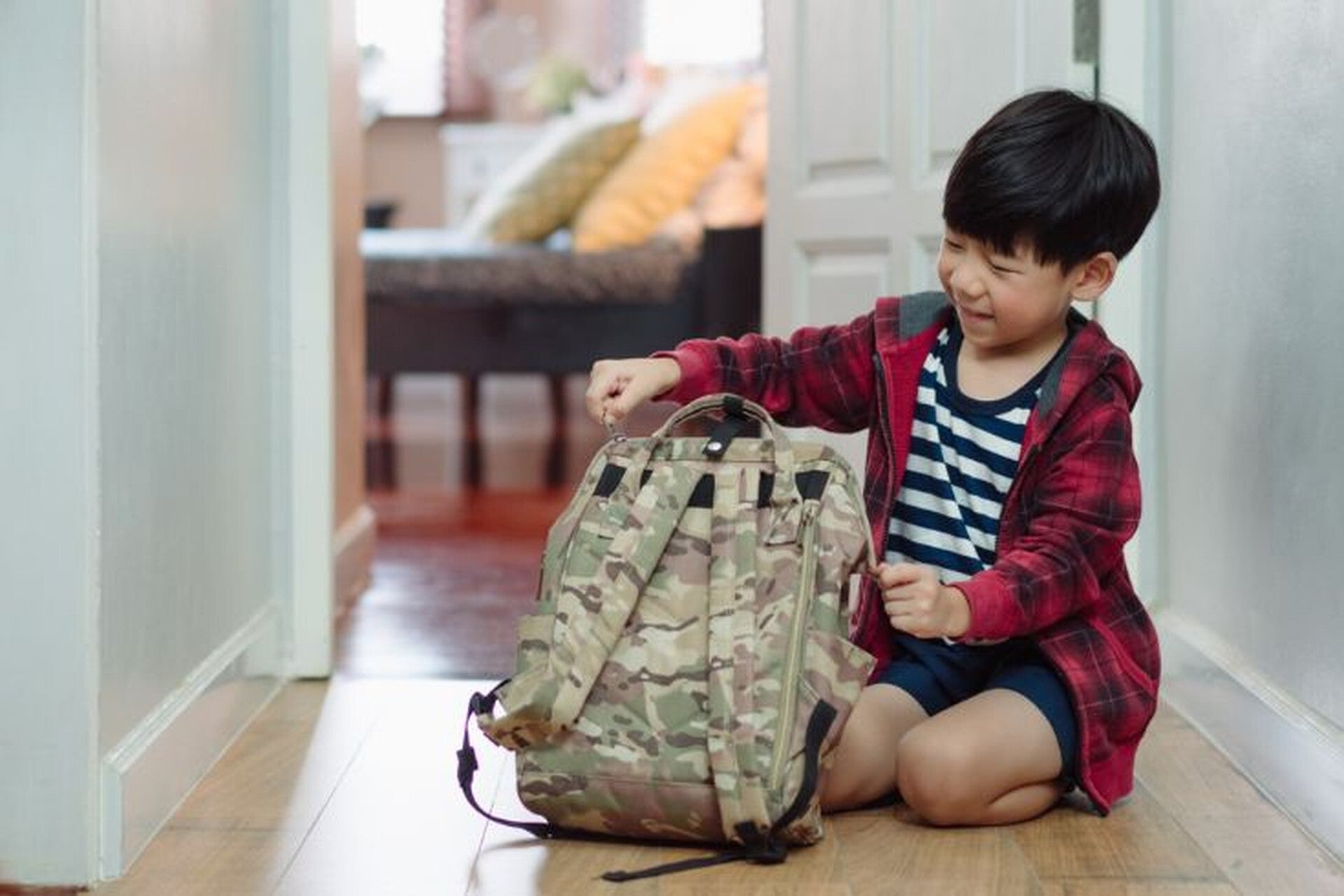 A young Asian preschooler closing his backpack while seated on the floor in the hallway.