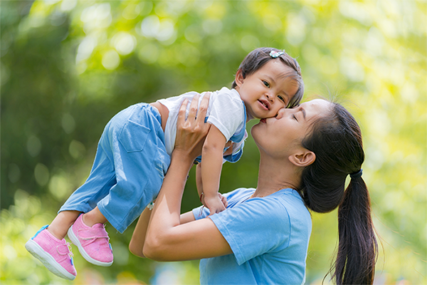 Mother lifting and kissing her toddler child outdoors in a sunlit park, candid moment of love and affection, family bonding and happy motherhood concept with soft green bokeh background.