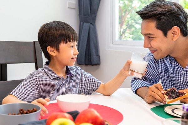 A happy young Asian boy having breakfast with his dad on Father’s Day and offering him a glass of milk in the morning.
