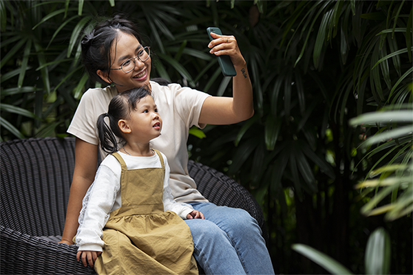 Mother and young daughter taking a selfie together on a bench outdoors, smiling and bonding in a lush garden setting, candid family moment with smartphone and natural greenery background.