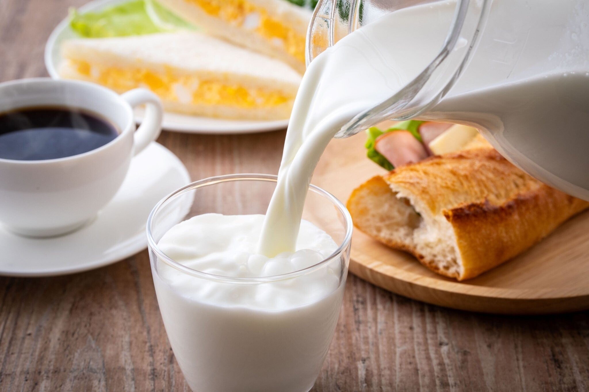 Milk from a jug being poured into a glass on a table set with breakfast food like egg and ham sandwiches and coffee. 