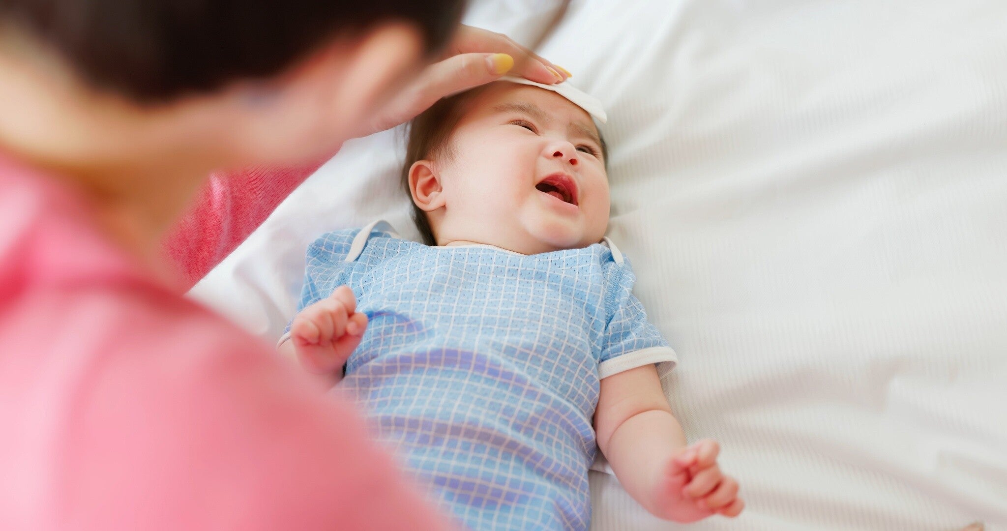 Photo of a fussy Asian baby with a fever while his mother checks his temperature on his forehead.
