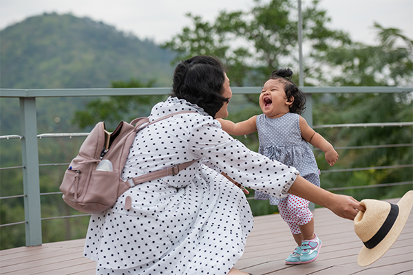 Mother and toddler laughing together on an outdoor deck with scenic mountain view, playful candid moment of family bonding, child reaching out while mom kneels with hat, joyful motherhood and travel lifestyle concept