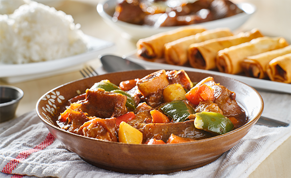 Photo of beef kaldereta in a bowl with plates of lumpiang Shanghai and white rice in the background