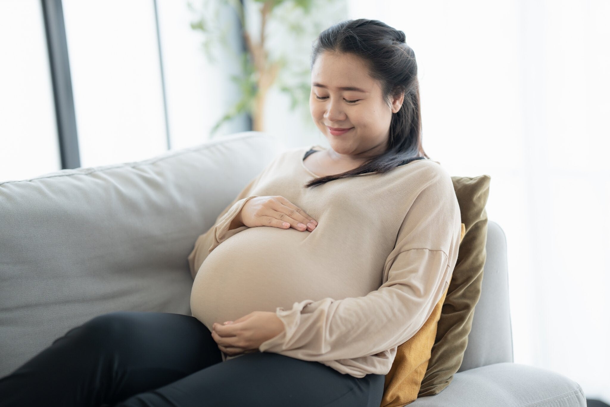 Pregnant Asian mom in cream, rubbing bump while on the couch, pondering about Filipino myths.