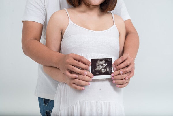 Pregnant Asian couple hugging and holding an ultrasound over her baby bump against a white background.