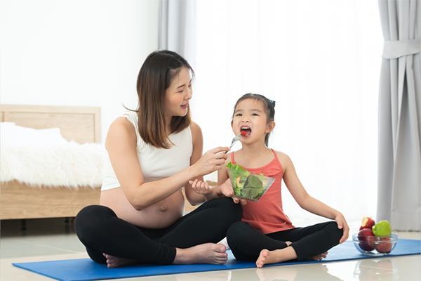 Pregnant Asian mom feeds her preschooler daughter vegetables after their yoga session