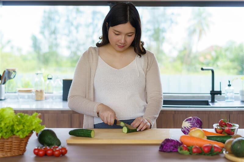 Pregnant Asian woman cutting cucumbers with vegetables in foreground.