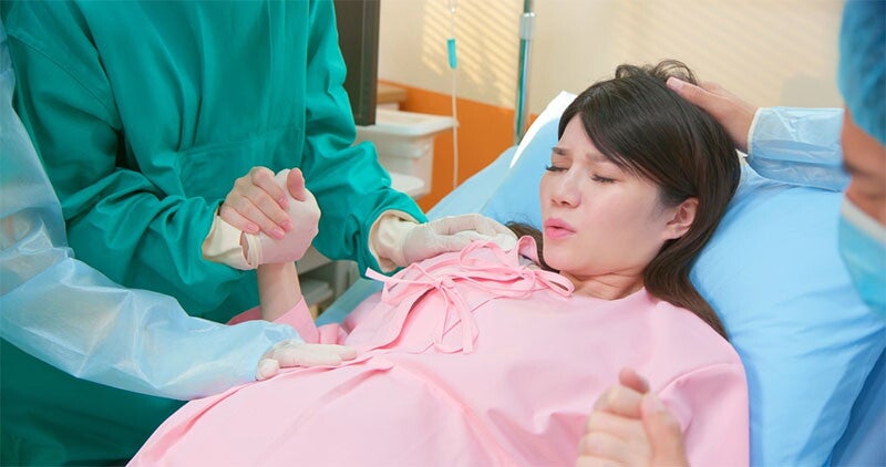 Pregnant Asian woman performs breathing exercise as she holds the hands of her partner and nurse during second stage of labor