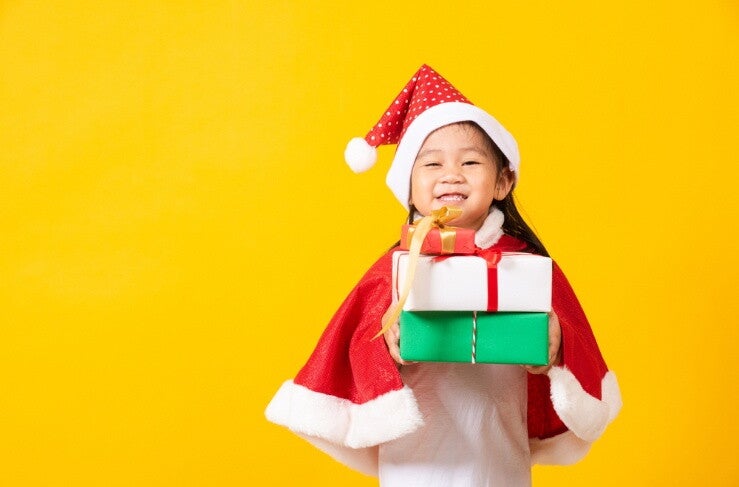 A smiling little Asian girl holding wrapped presents and wearing a Santa hat and cape against yellow background.