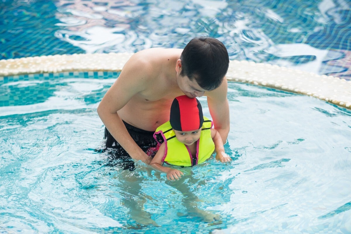 Little Asian girl wearing life jacket in the pool, swimming together with her dad. 