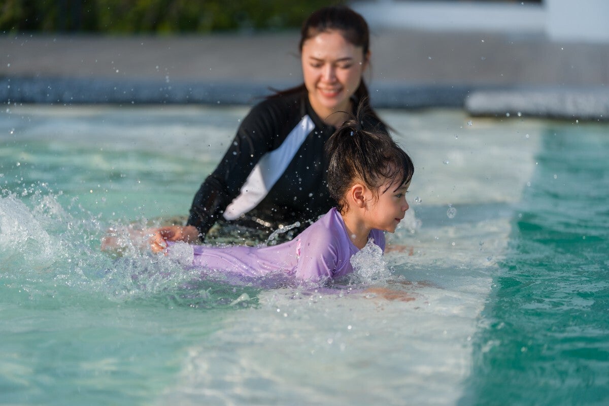 Young Asian mother with her preschool girl kicking water while practicing swimming at the edge of a pool.