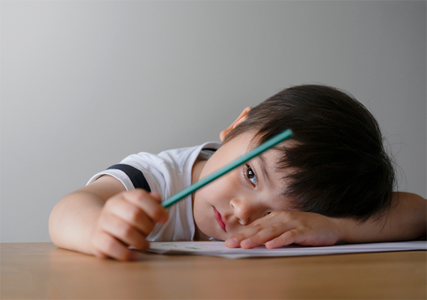 Sad young preschooler with his head on his school desk stares into space while holding a pencil