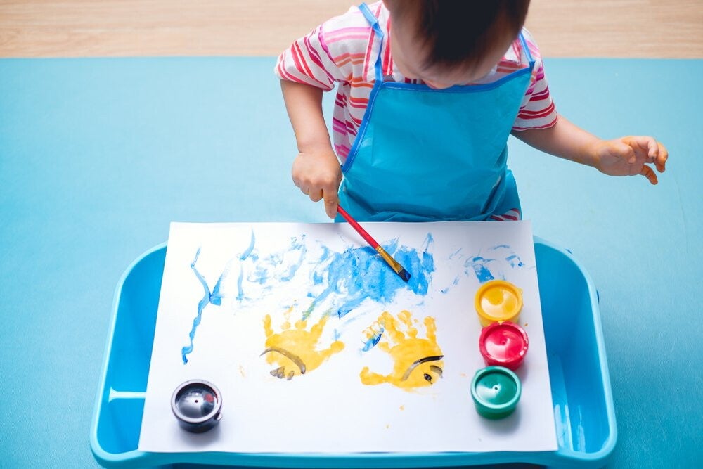 Asian toddler uses a brush to paint on paper with yellow handprints. 