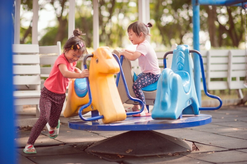 Two healthy Asian female children of different ages play on a merry-go-round at a park.