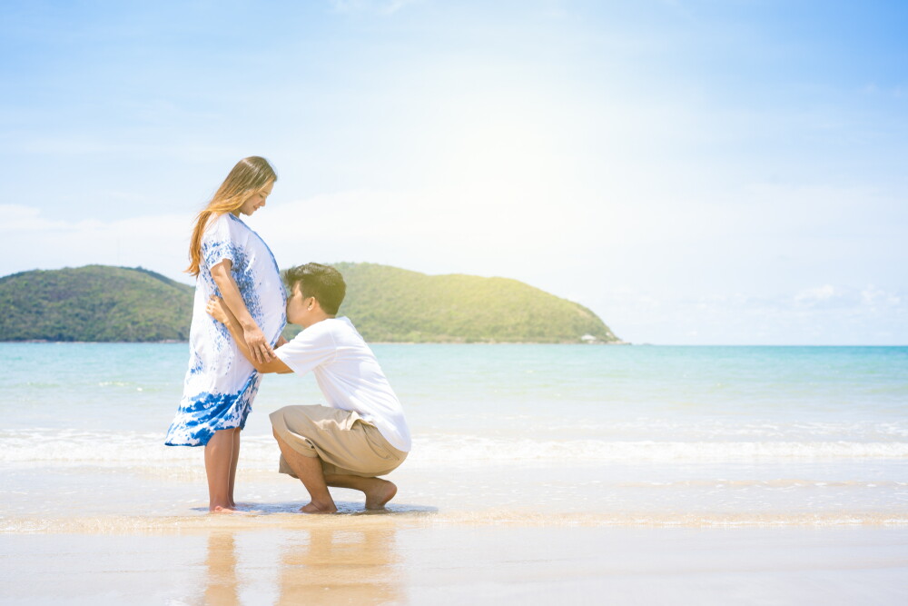 Asian man squats to kiss his pregnant partner’s baby bump while they’re at a beach. 