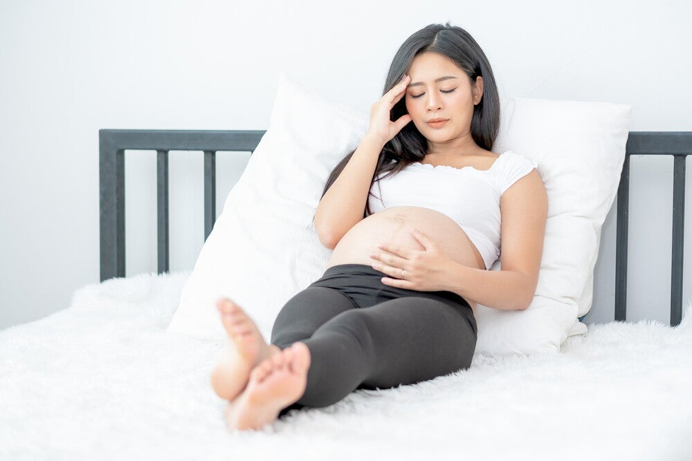  A young pregnant Asian woman with a hand on her temple and one on her belly, resting in bed.