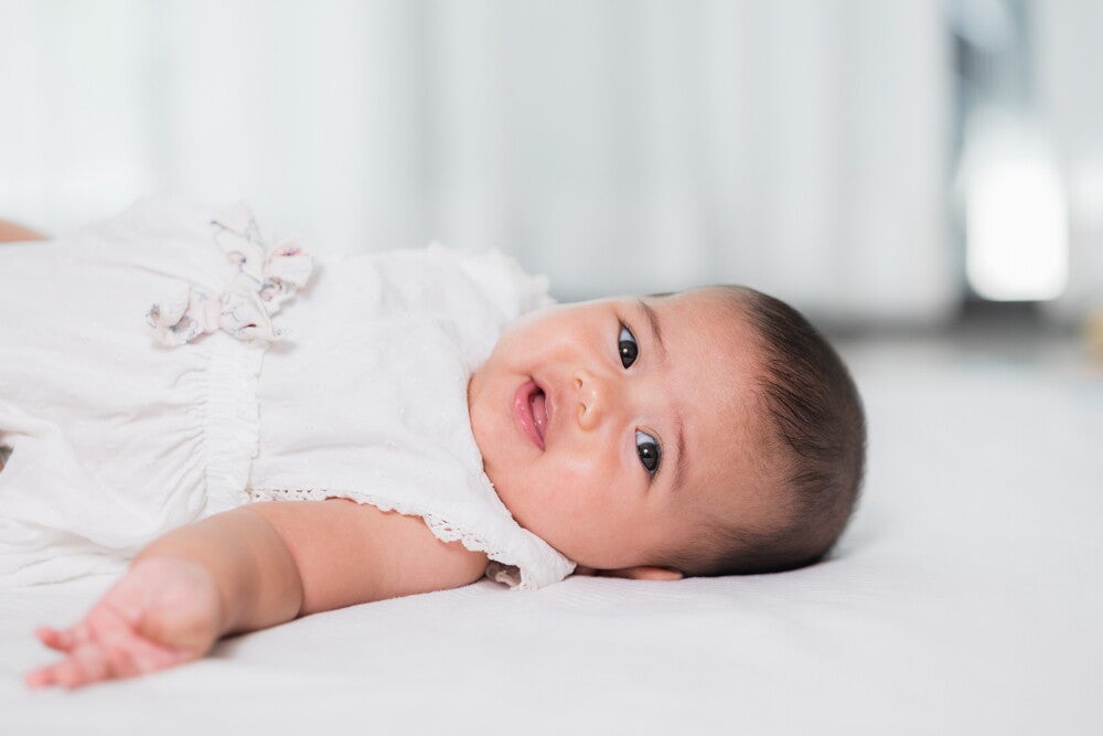Adorable Asian baby in her white christening outfit lies in bed.