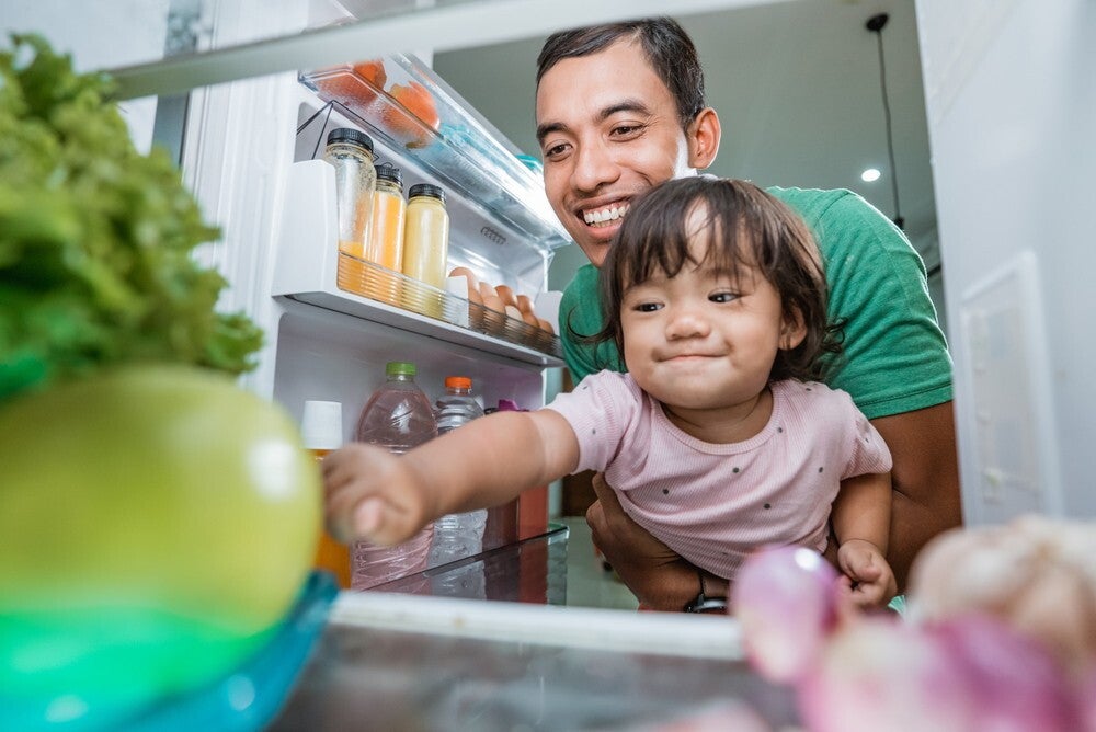 Female Asian toddler reaches for a fruit in the refrigerator as her dad carries her.
