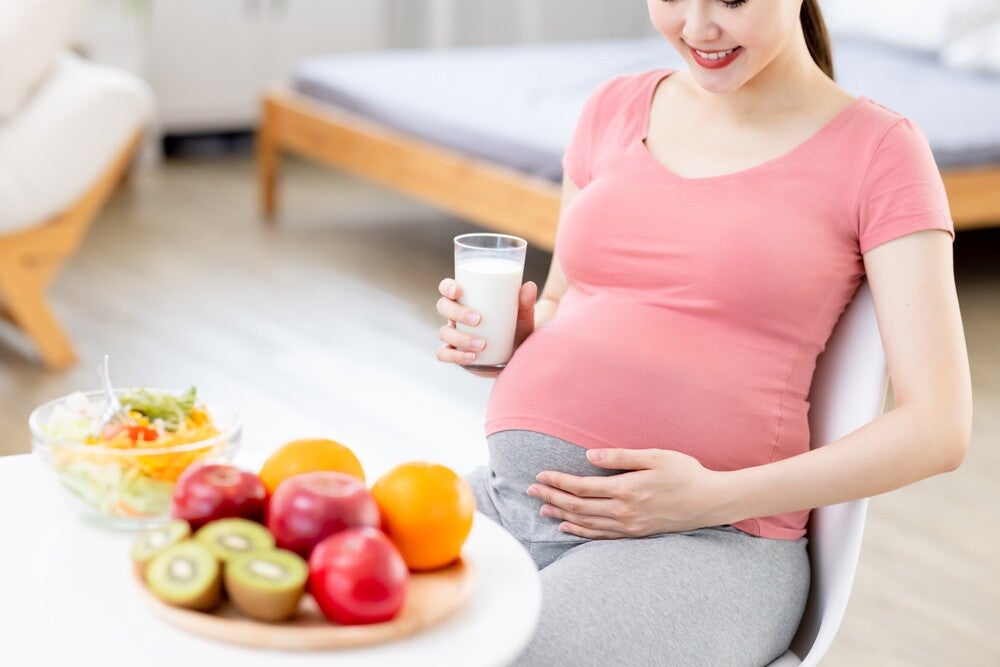 Asian pregnant woman smiles at her baby bump, holding a glass of milk and fruits in the foreground.