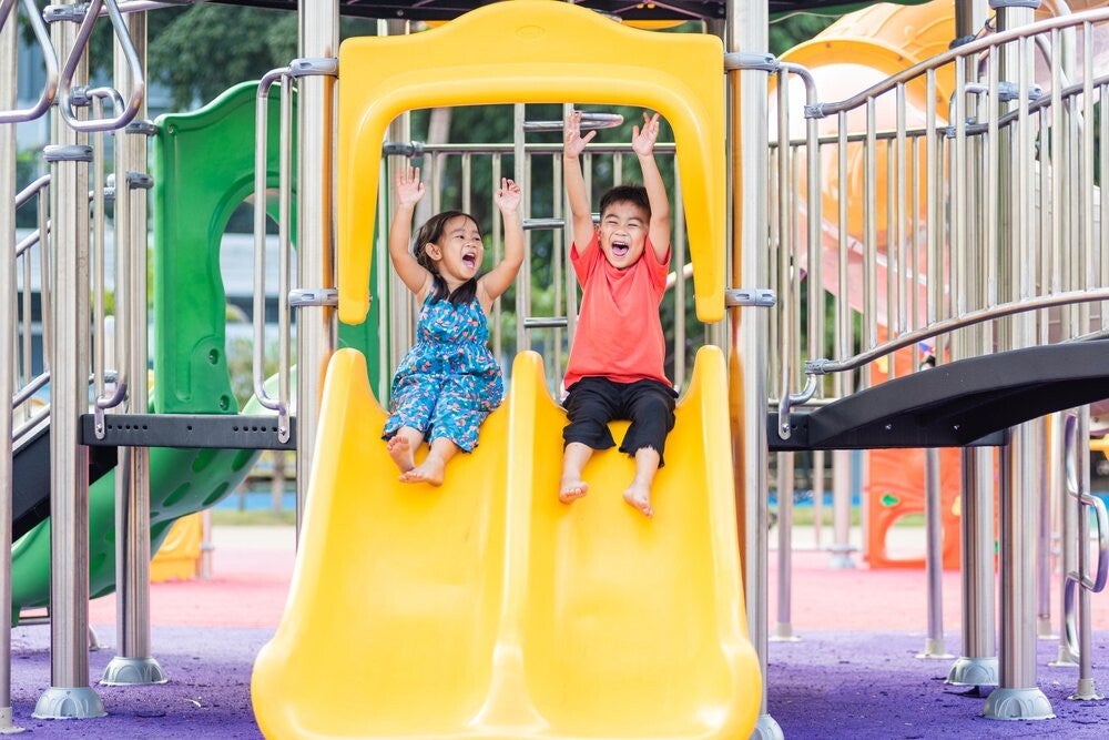 Two Asian children lift their arms as they prepare to go sliding at a playground.