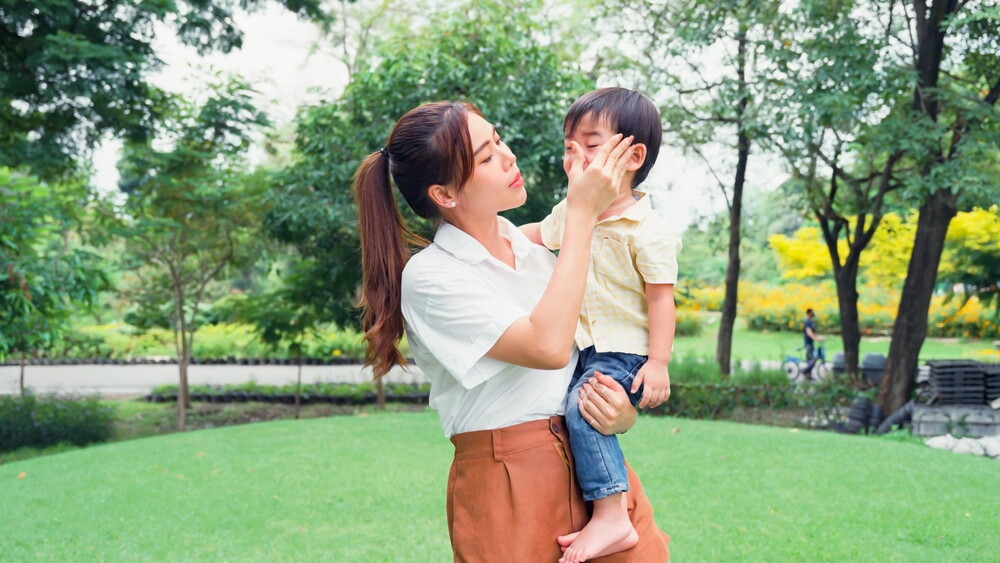 Asian mom practicing gentle parenting wipes her son’s tears as she carries him at the park.