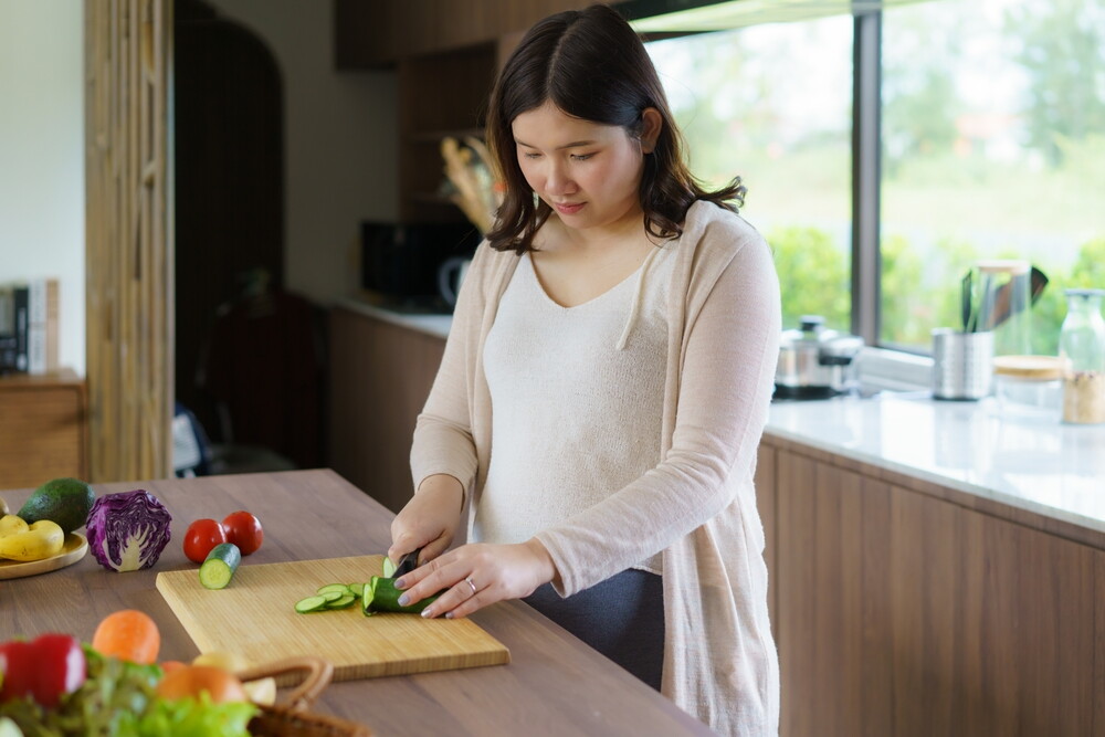 Asian pregnant woman cuts cucumber as she prepares a healthy meal in the kitchen.