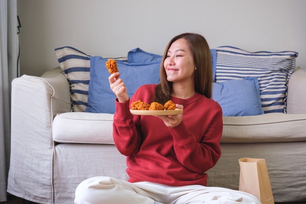 Young Asian woman smiles as she looks at the fried chicken leg she's holding aloft from a plate.