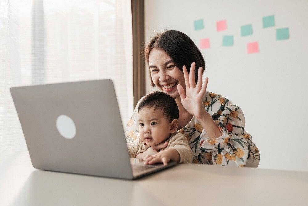 Asian work-from-home mom puts her baby on her lap as she conducts a virtual meeting on her laptop.