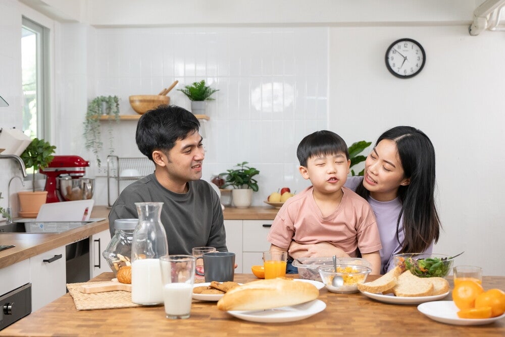 A family sits together at a kitchen table enjoying breakfast, with various foods such as bread, milk, juice, fruit, and salad laid out in front of them. The kitchen in the background features white tiles, wooden countertops, plants, and a wall clock.