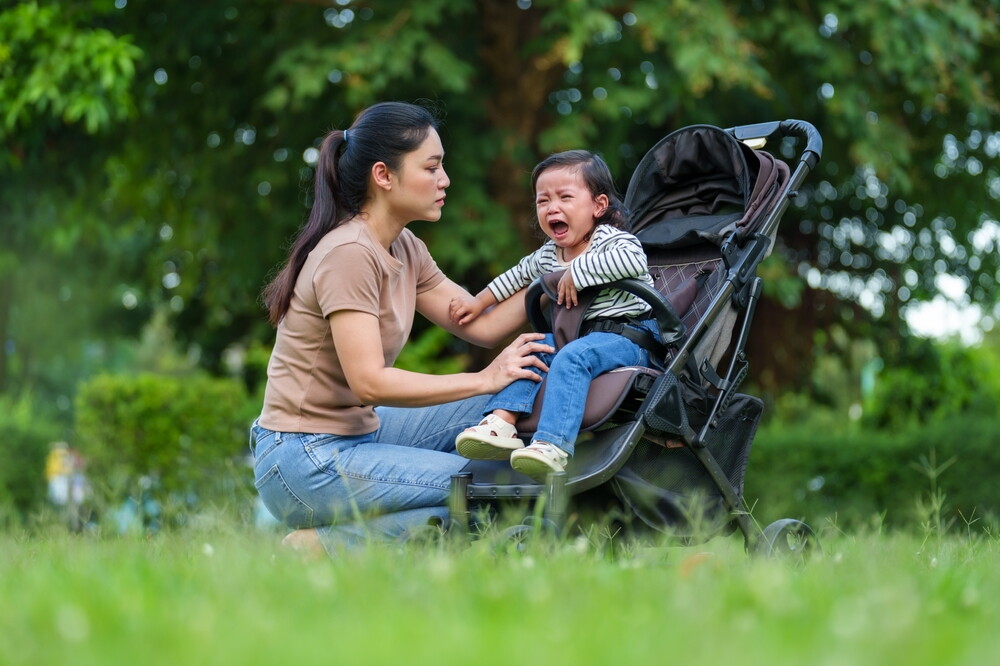 Asian mom squats to talk at eye level to her crying toddler sitting in the stroller.