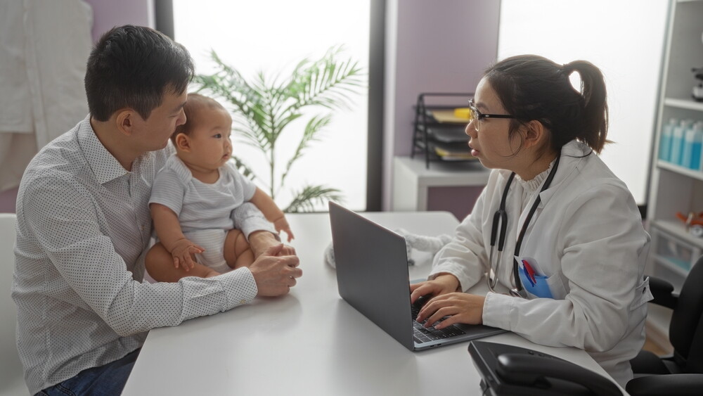 Asian dad, with his baby seated on the desk, talks to a female doctor working on her laptop.